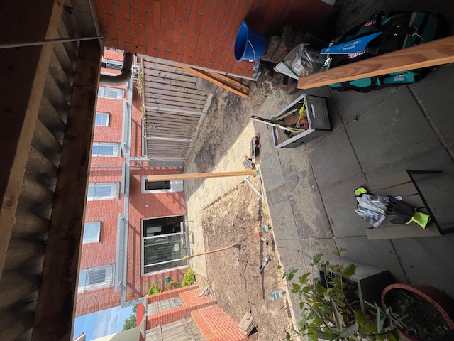 Achtertuin met kale grond langs een bakstenen muur en houten schutting tijdens de aanleg
